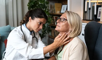 Female doctor examining an older woman’s neck and throat area during a home visit. The doctor appears focused and attentive, while the patient sits calmly in a calmly decorated office.